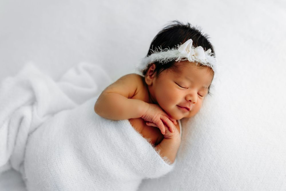 Peaceful sleeping newborn wrapped in white blanket wearing delicate lace headband during professional portrait session.
Rashinphotography-