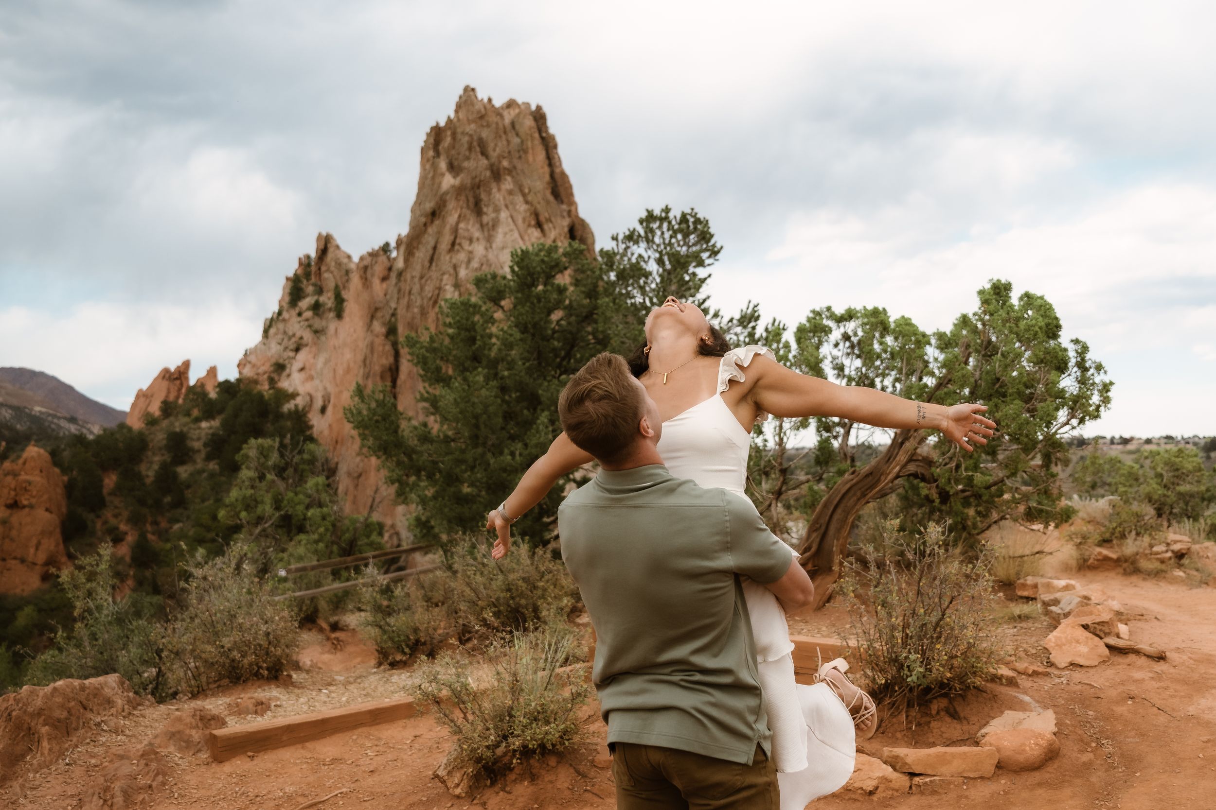 A couple poses playfully against dramatic red rock formations in an outdoor setting.