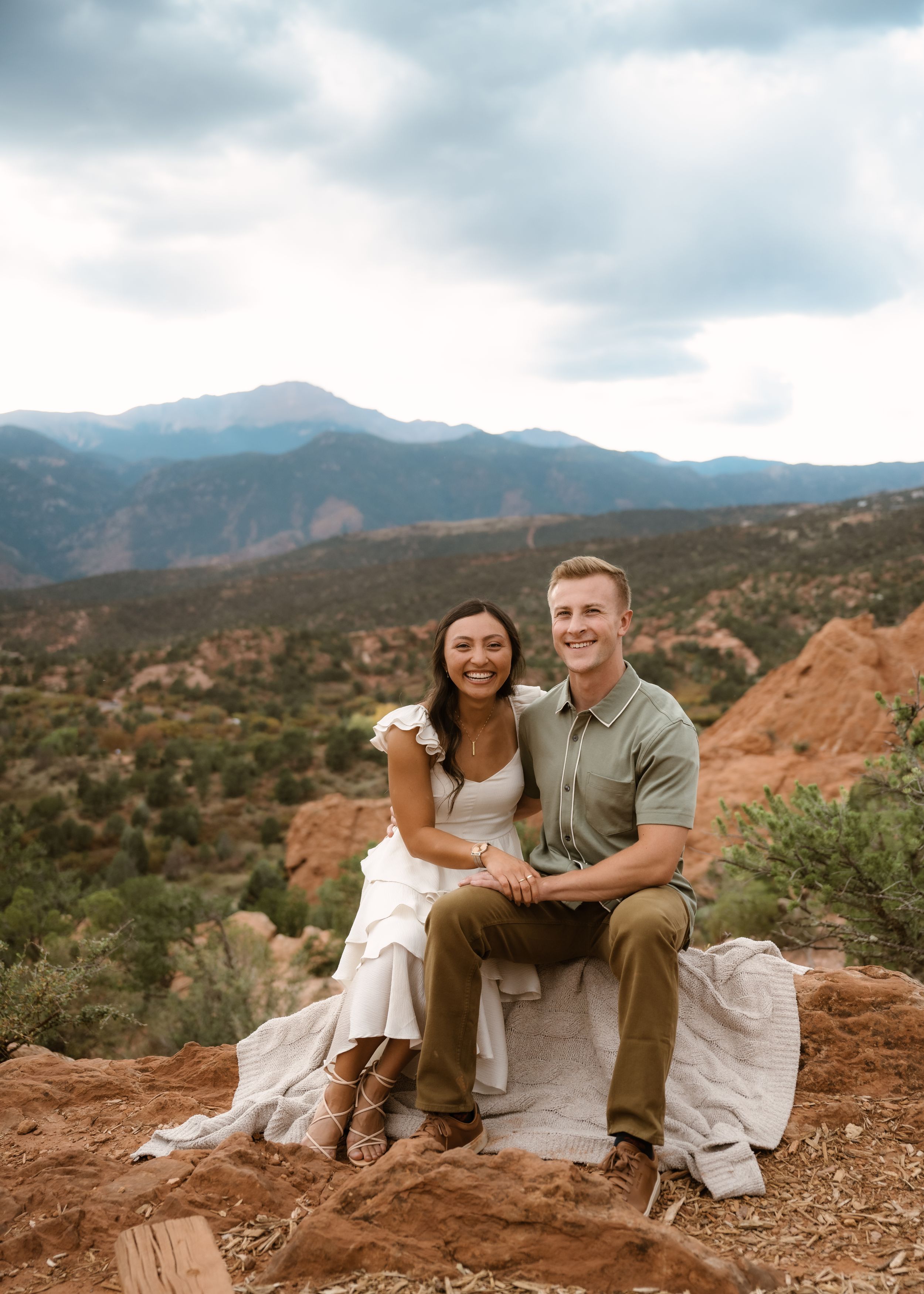 A romantic couple sits together on red rocks with mountains in the background at Garden of the Gods in Colorado.