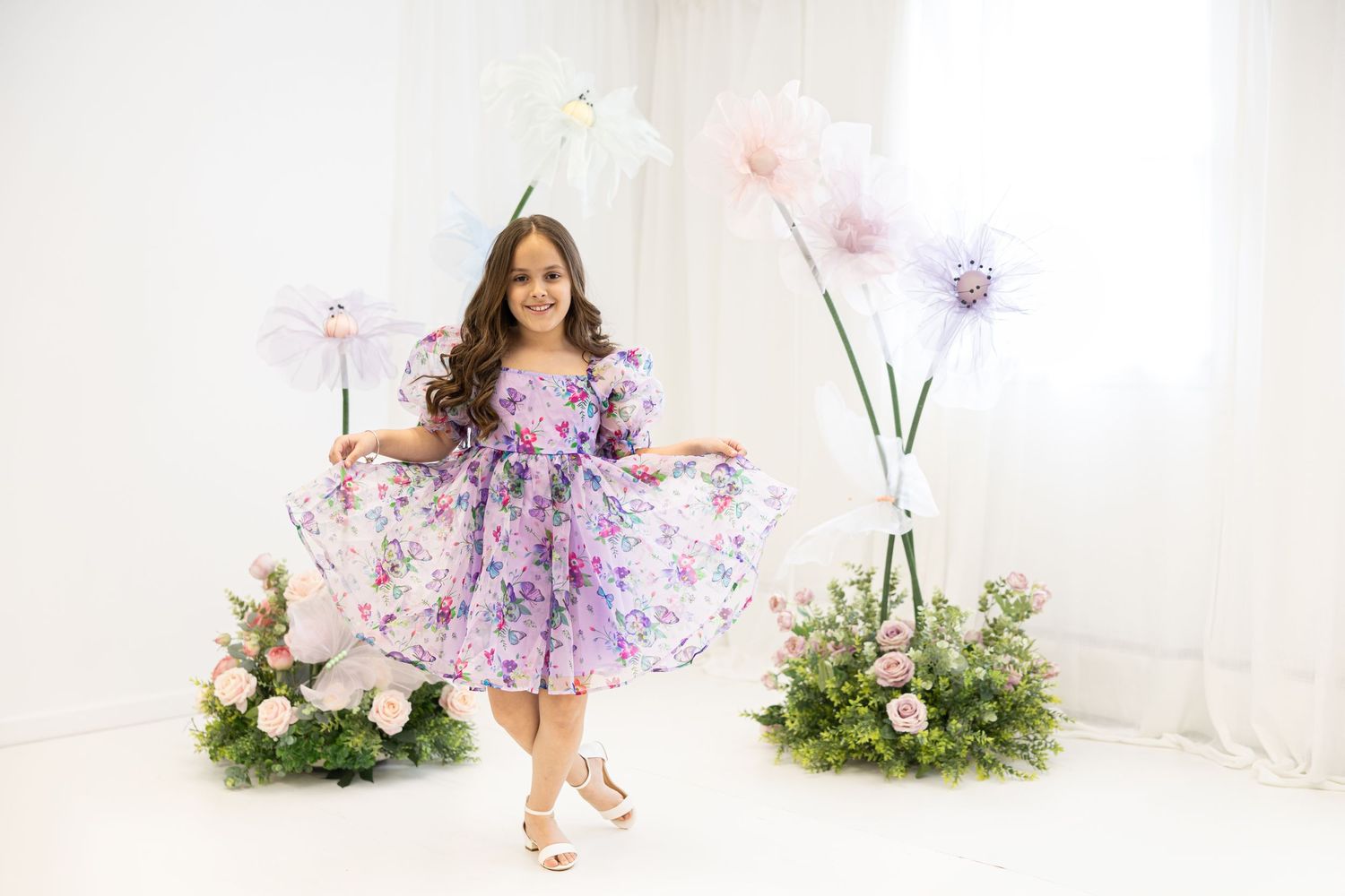 Child in purple floral dress twirls among garden-themed studio setup with giant flower decorations.