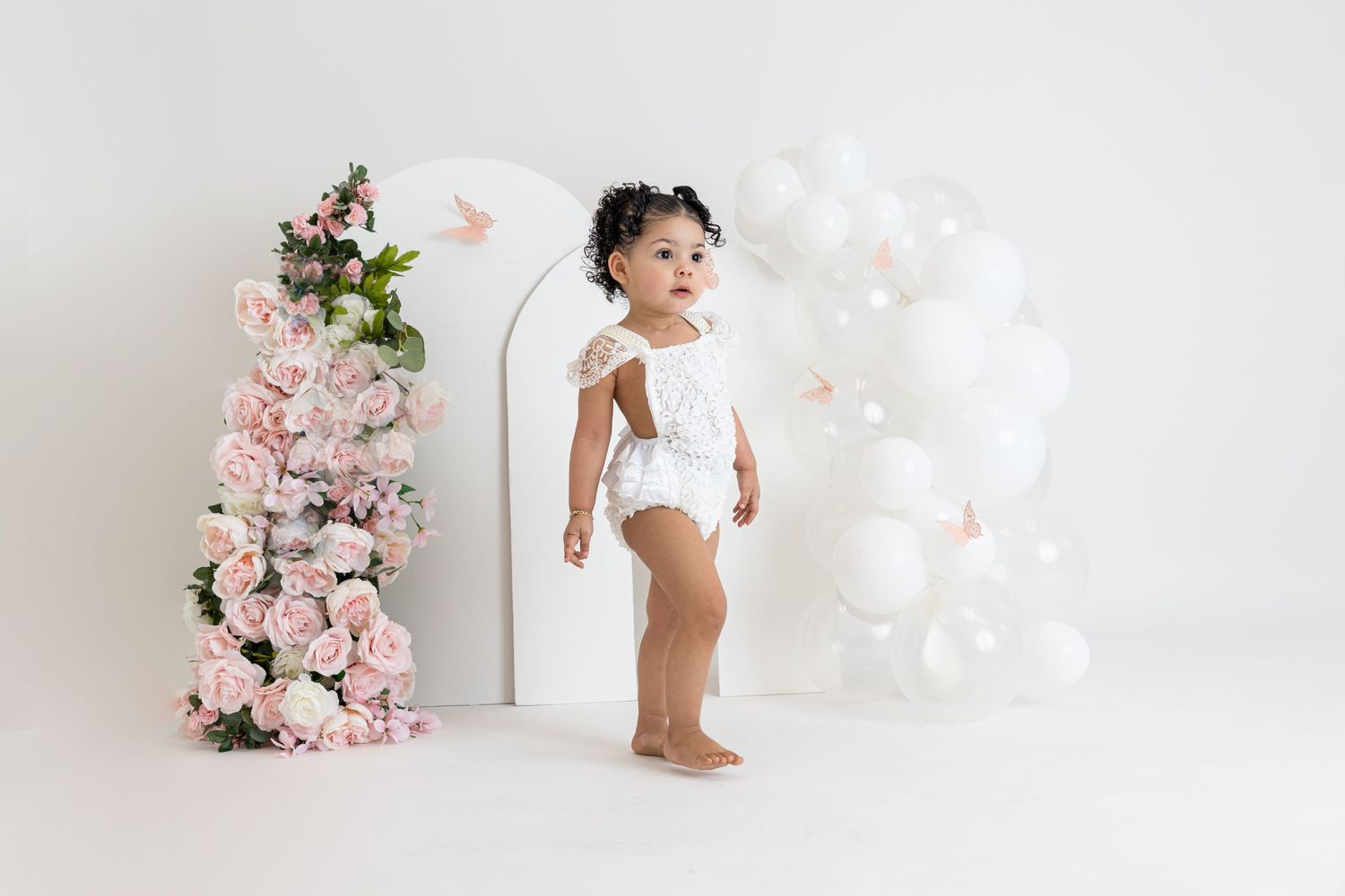 Small child in white lace romper poses near pink floral arch and white balloon decorations.