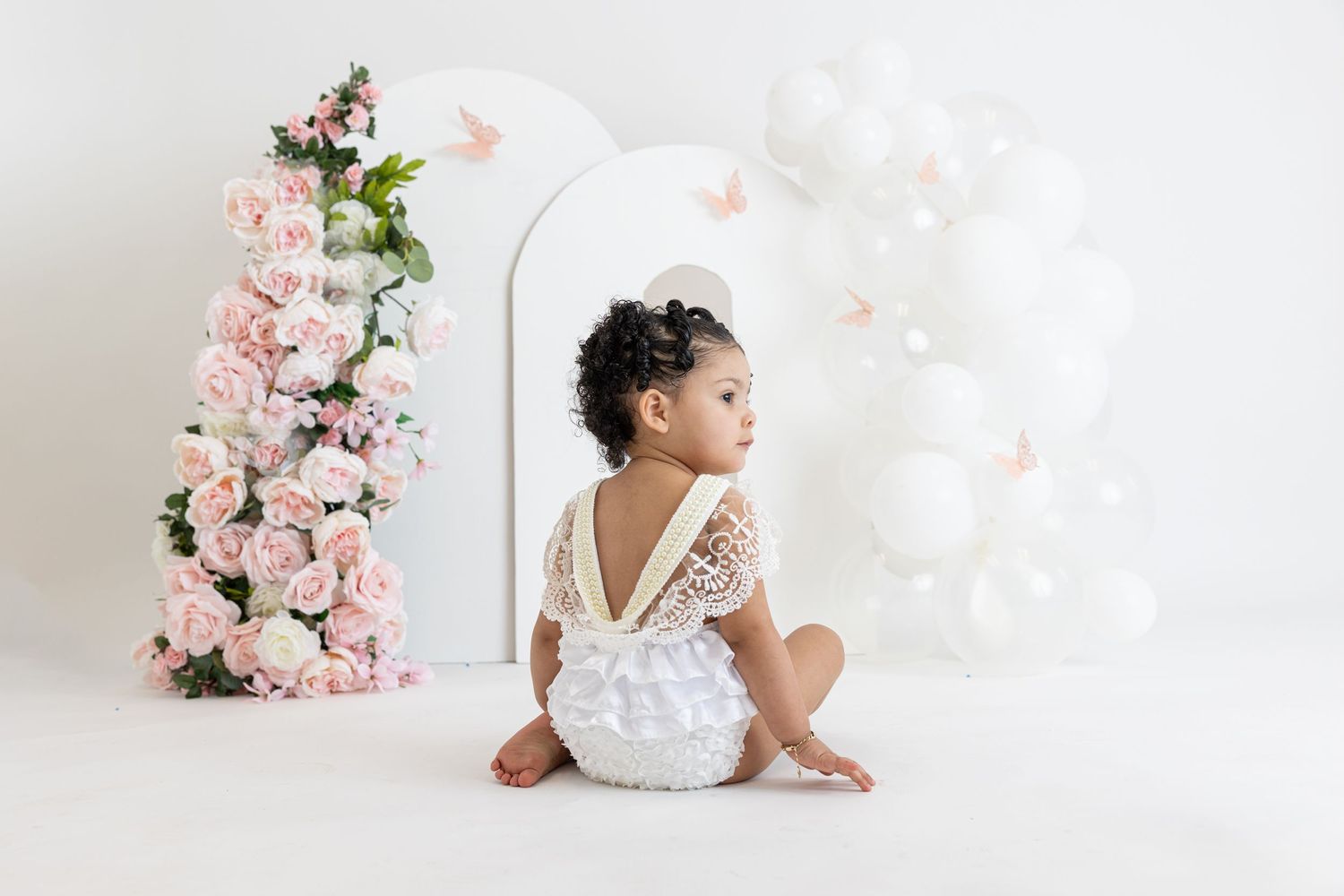 Small child in white outfit sits on floor near pink floral decorations and white balloons.