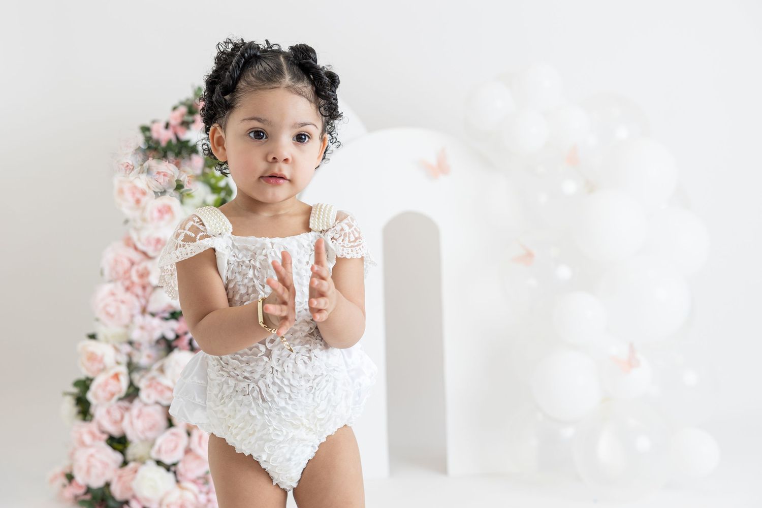 Small child wearing white lace outfit poses in front of pink floral arch and white balloon backdrop.