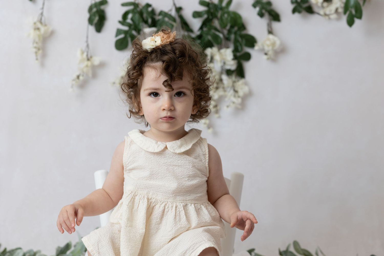 Small child in cream dress sits against white background with hanging green foliage and flowers.