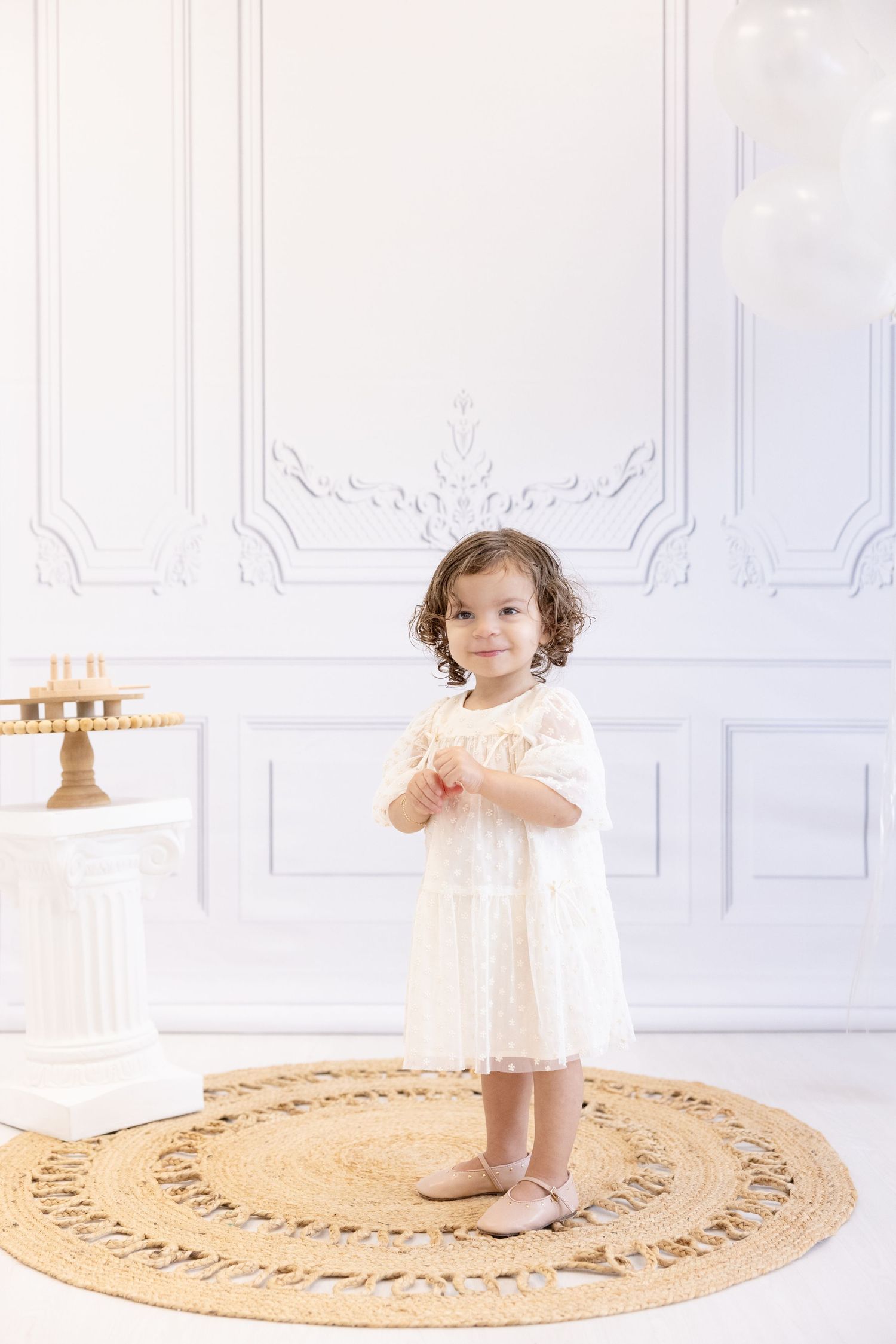 Child in white dress stands on round rug in elegant white room setting.
