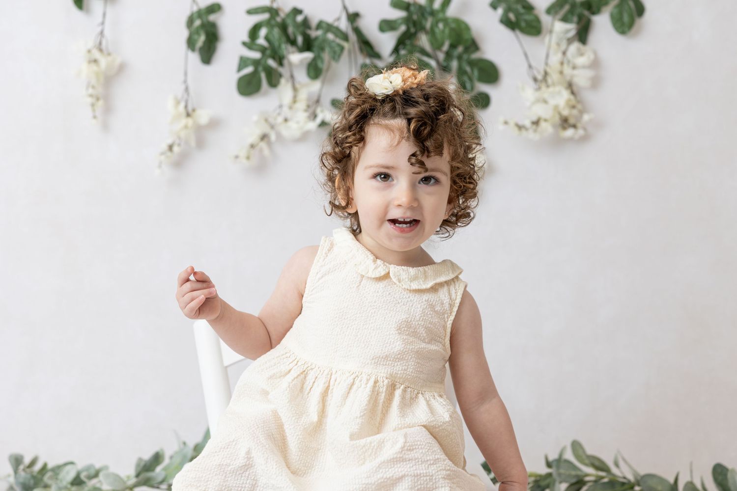 Toddler in cream dress smiles while sitting against botanical wall decor.