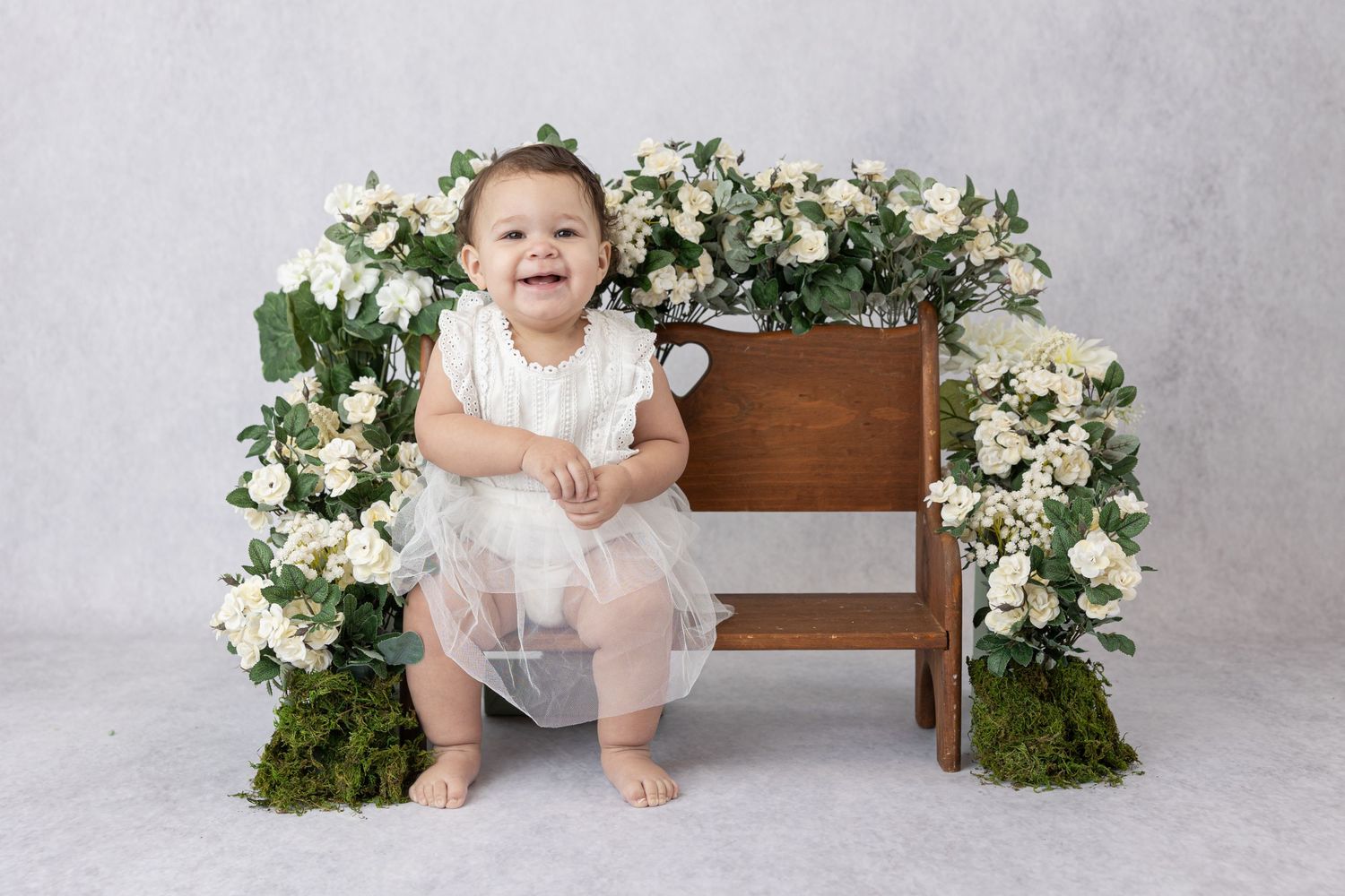 Baby in white dress sitting by wooden chair decorated with white floral arrangements.