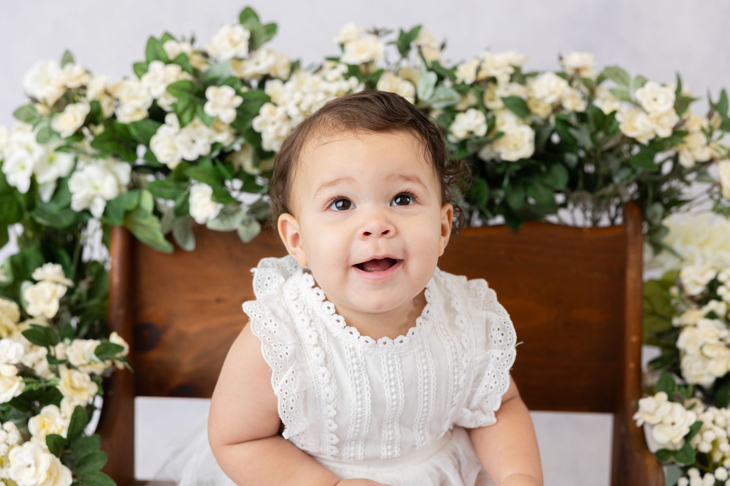 Baby in ruffled white dress smiling in front of wooden chair with floral decorations.