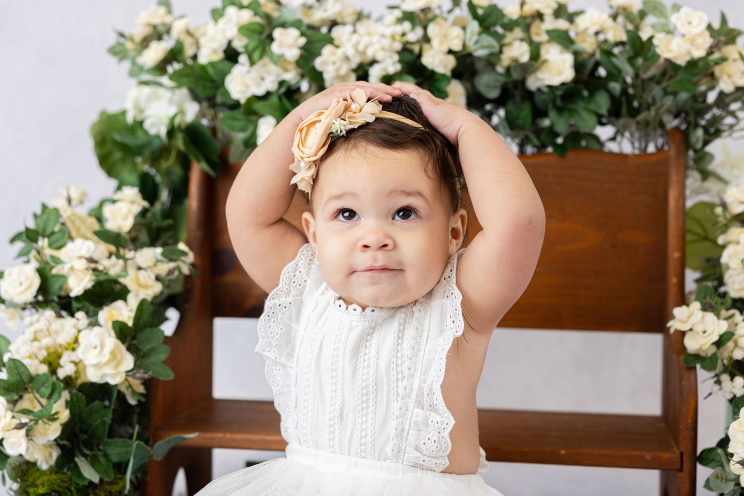 Baby in white dress touching head while sitting against floral backdrop.