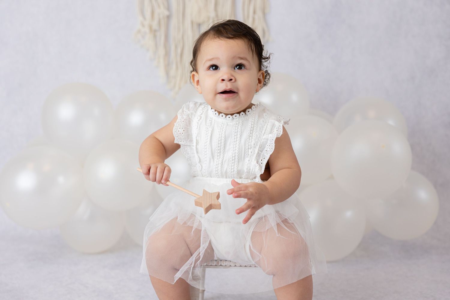 Baby in white tulle outfit sitting among white balloons in studio setting.
