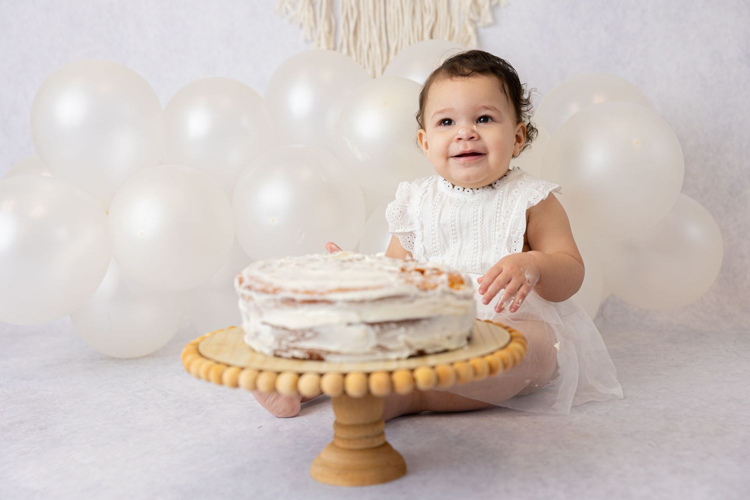 Baby in white dress reaching for cake on wooden stand with white balloon backdrop.