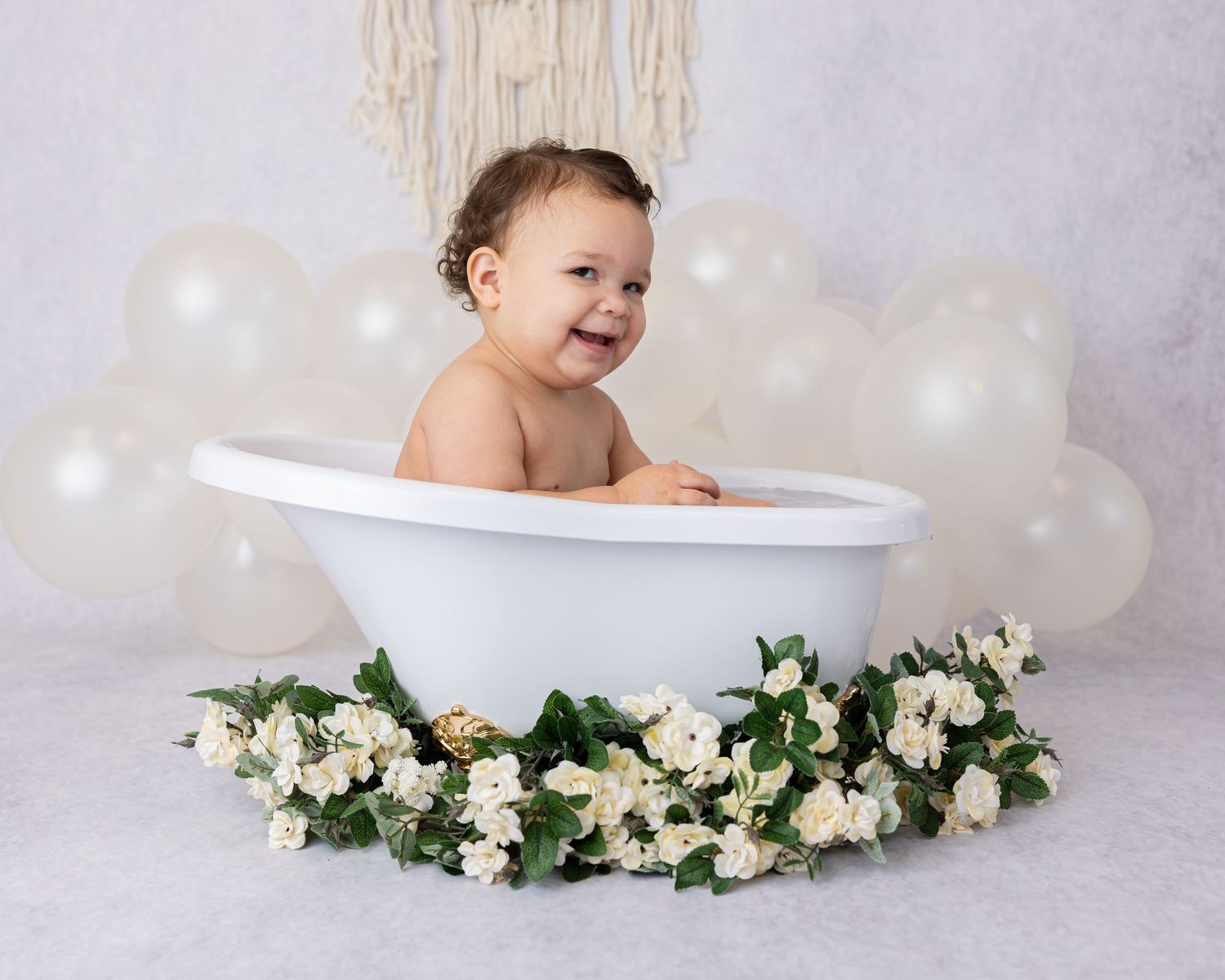 Baby sitting in decorative bathtub surrounded by white flowers and balloons.