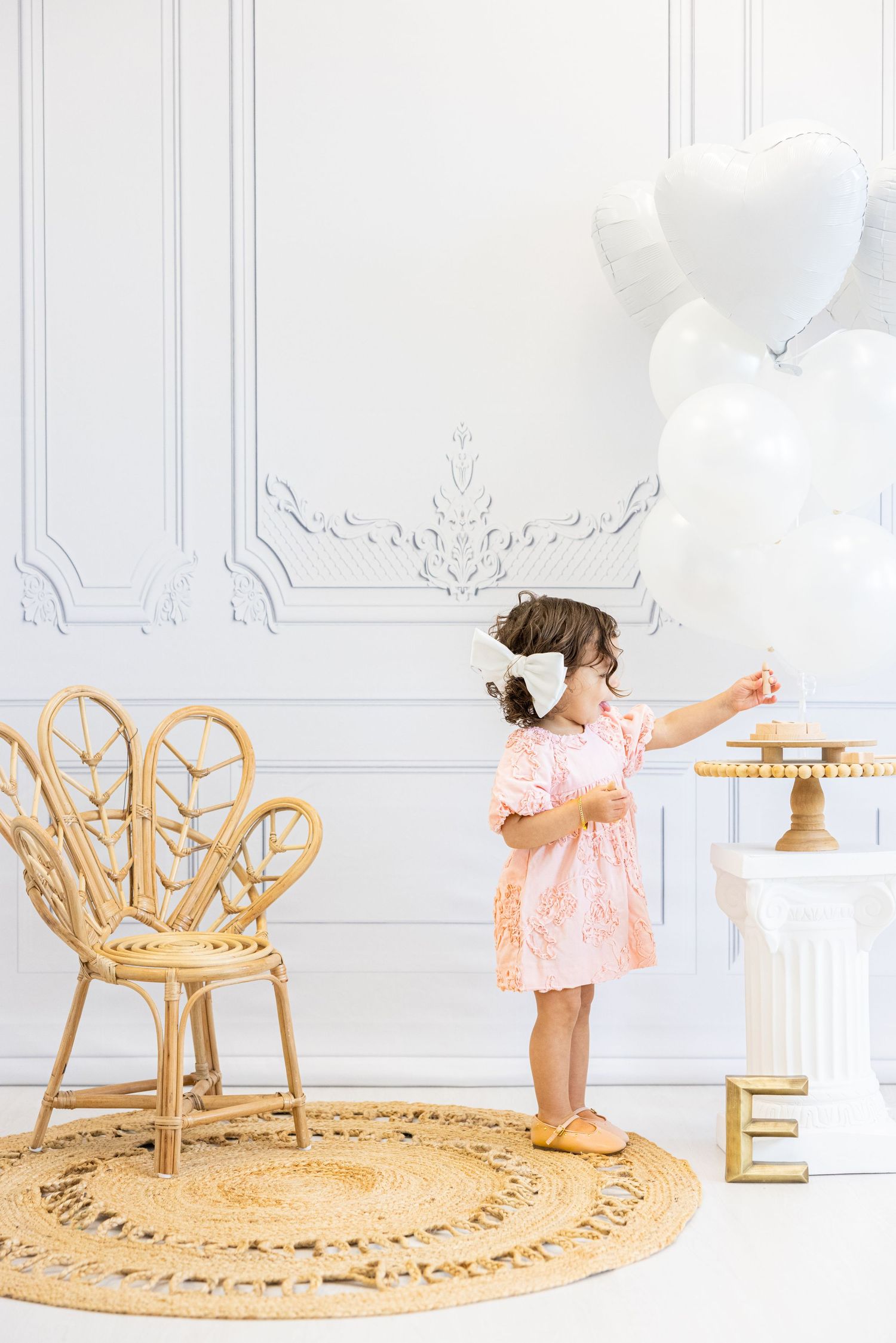 Young child in pink dress plays with white balloons beside rattan peacock chair in elegant white room.