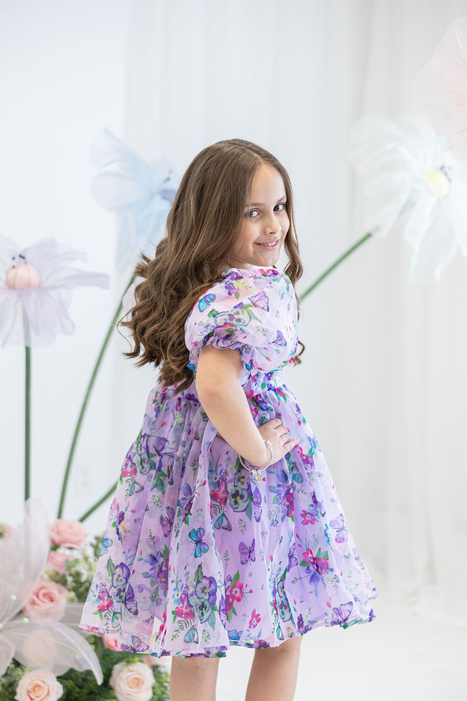 Child in purple floral dress poses among pastel flower decorations in white studio setting.