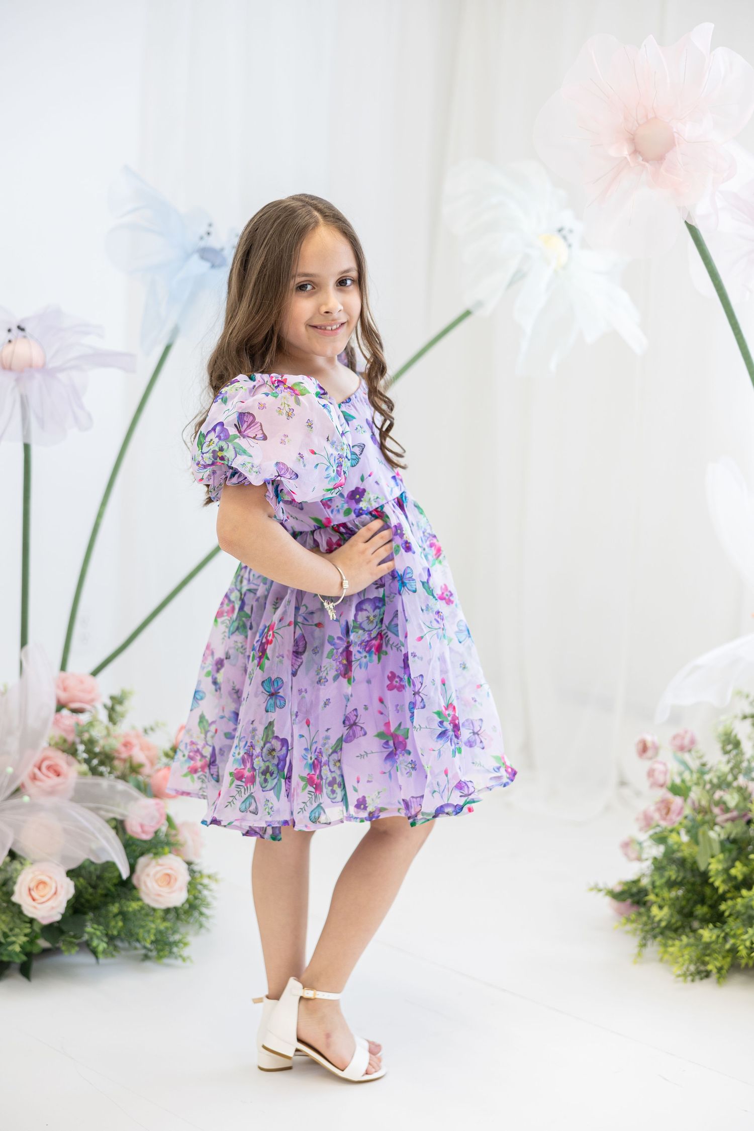 Child in a purple floral dress poses among pink roses and decorative flowers in a studio setting.