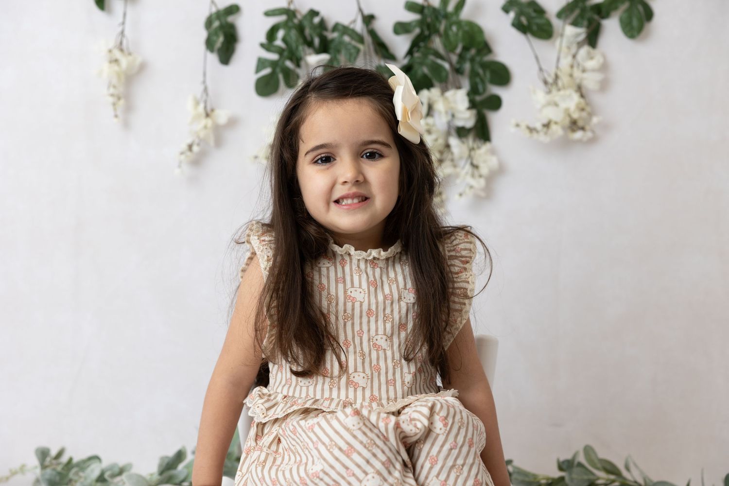 Child in striped dress poses in front of white wall decorated with hanging greenery and flowers.