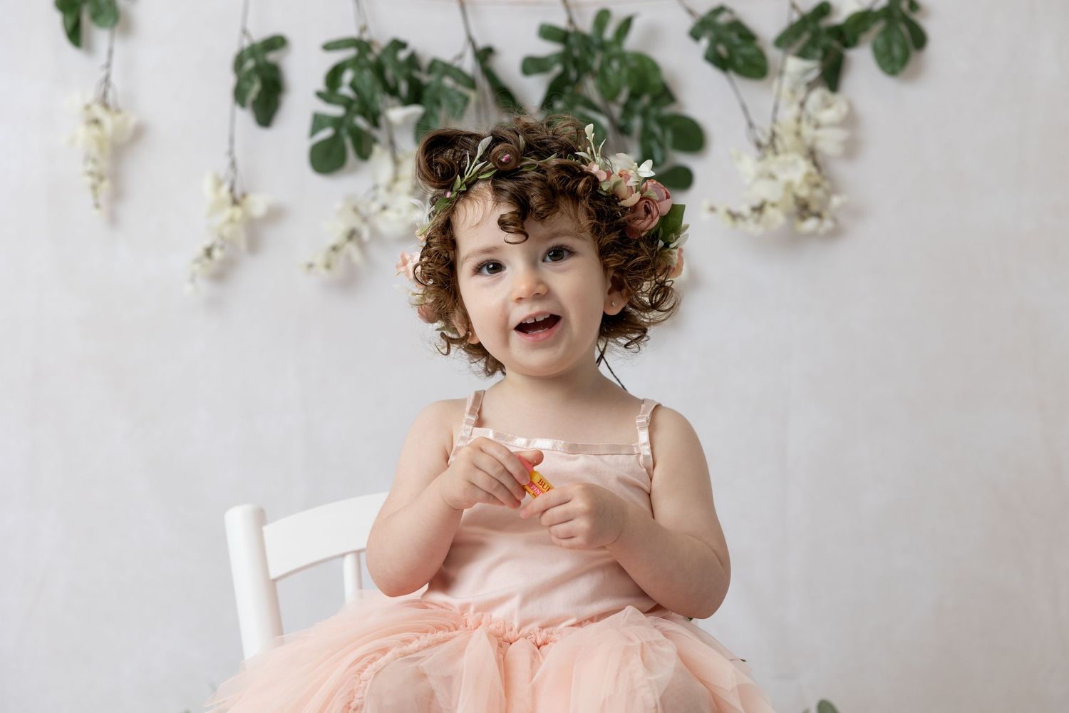 Child in peach tulle dress sits on white chair against floral wall backdrop.