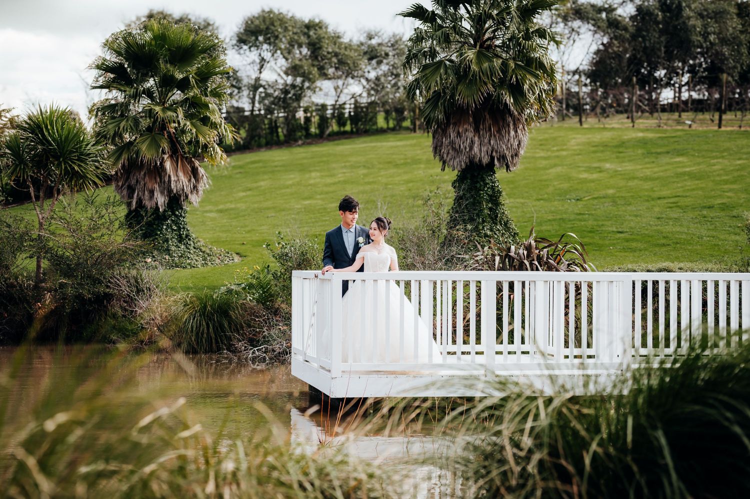Wedding couple stands on white bridge surrounded by palm trees and lush green landscape.