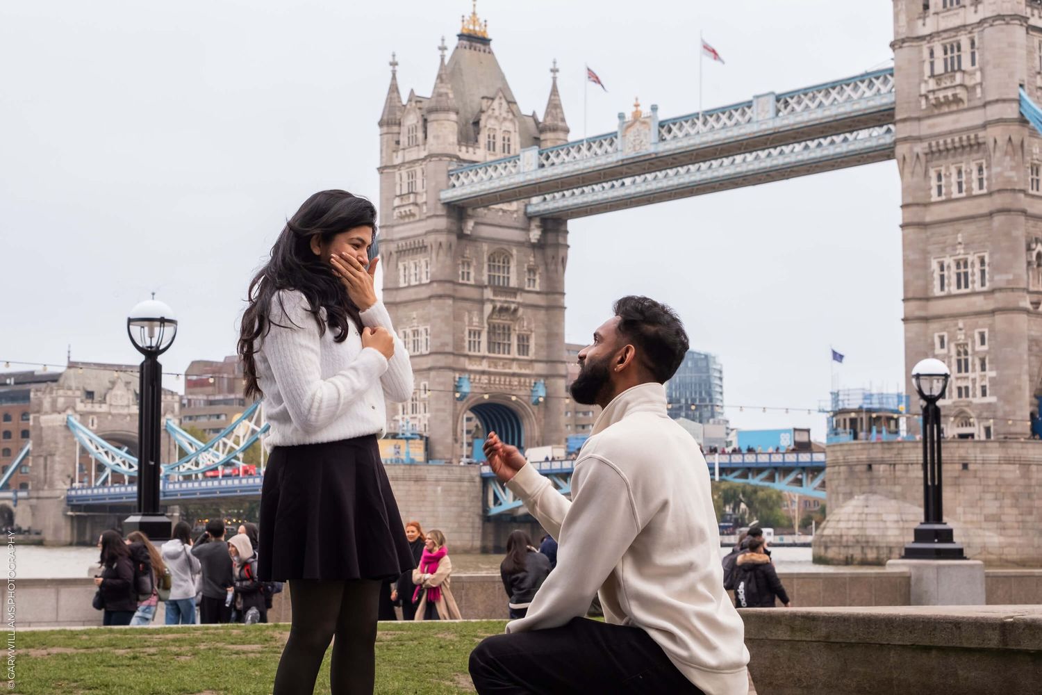 Romantic proposal scene near London's Tower Bridge on a cloudy day.