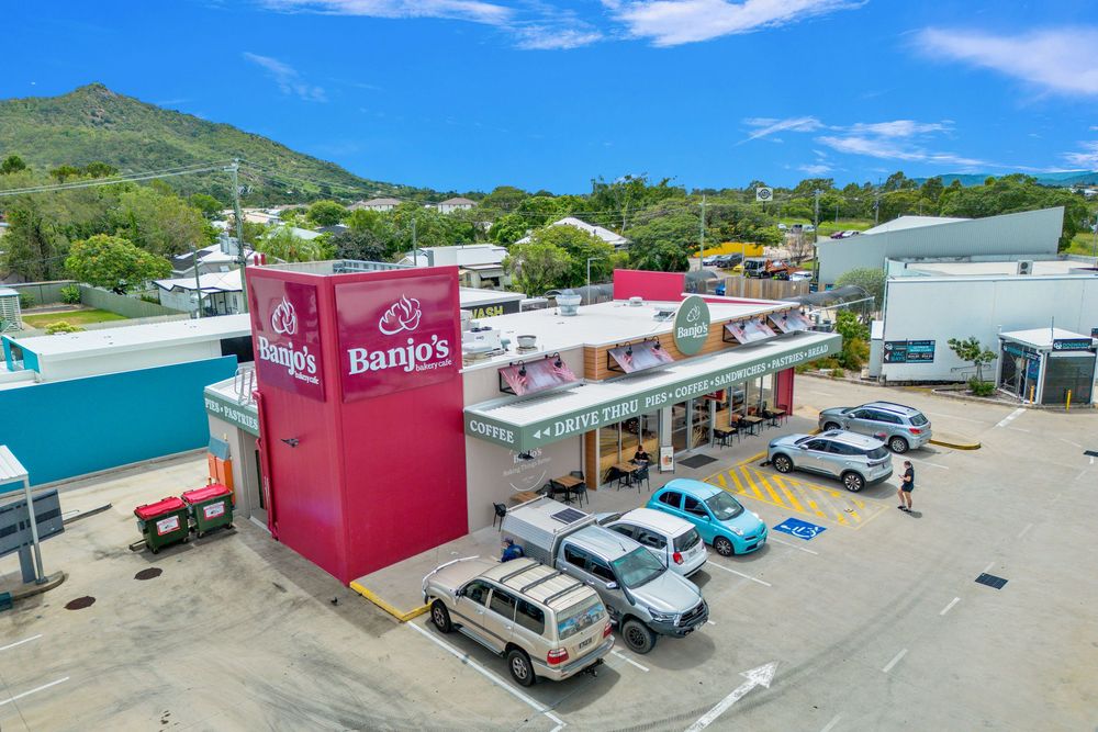 Aerial view of a coastal strip mall with parked cars and red business signage under bright blue skies.