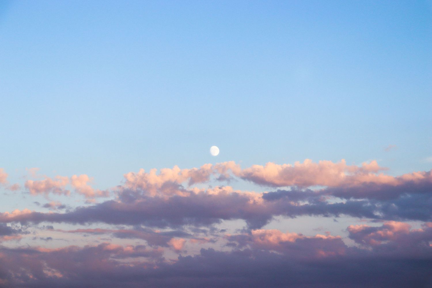 Moon rises through pink and purple sunset clouds in evening sky.