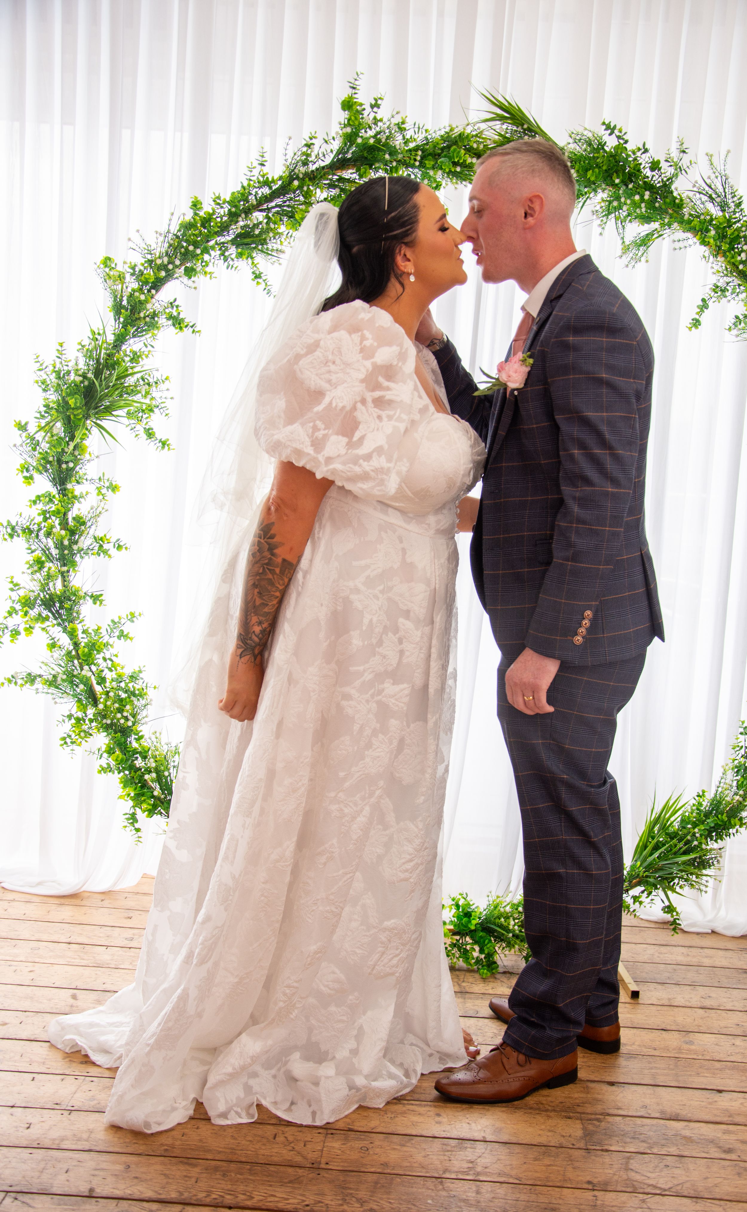 Romantic wedding kiss under a leafy green arch with white curtain backdrop.