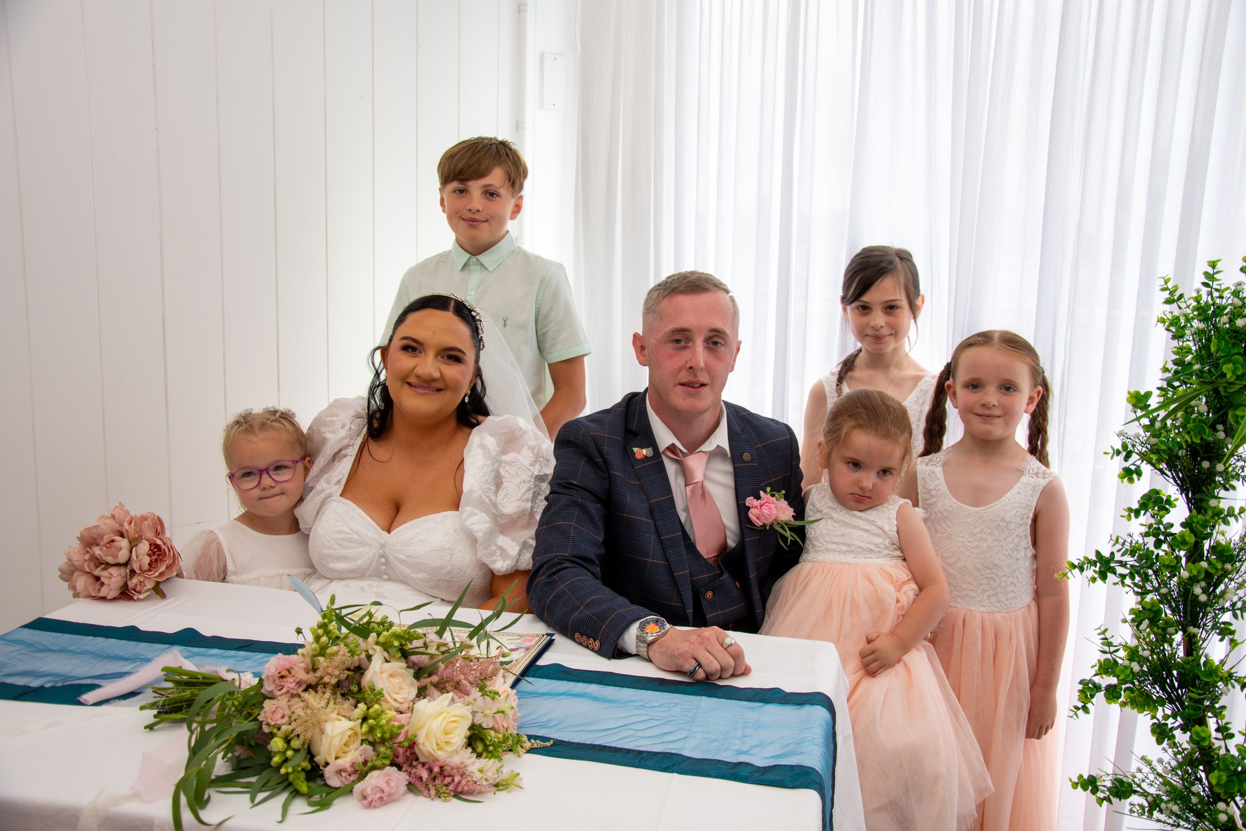 Wedding party gathers around signing table with floral decorations.