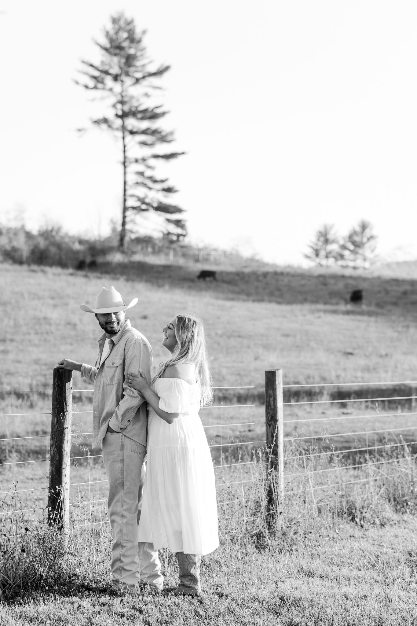 A black and white countryside wedding photo with a tall pine tree and rustic fence in the background.