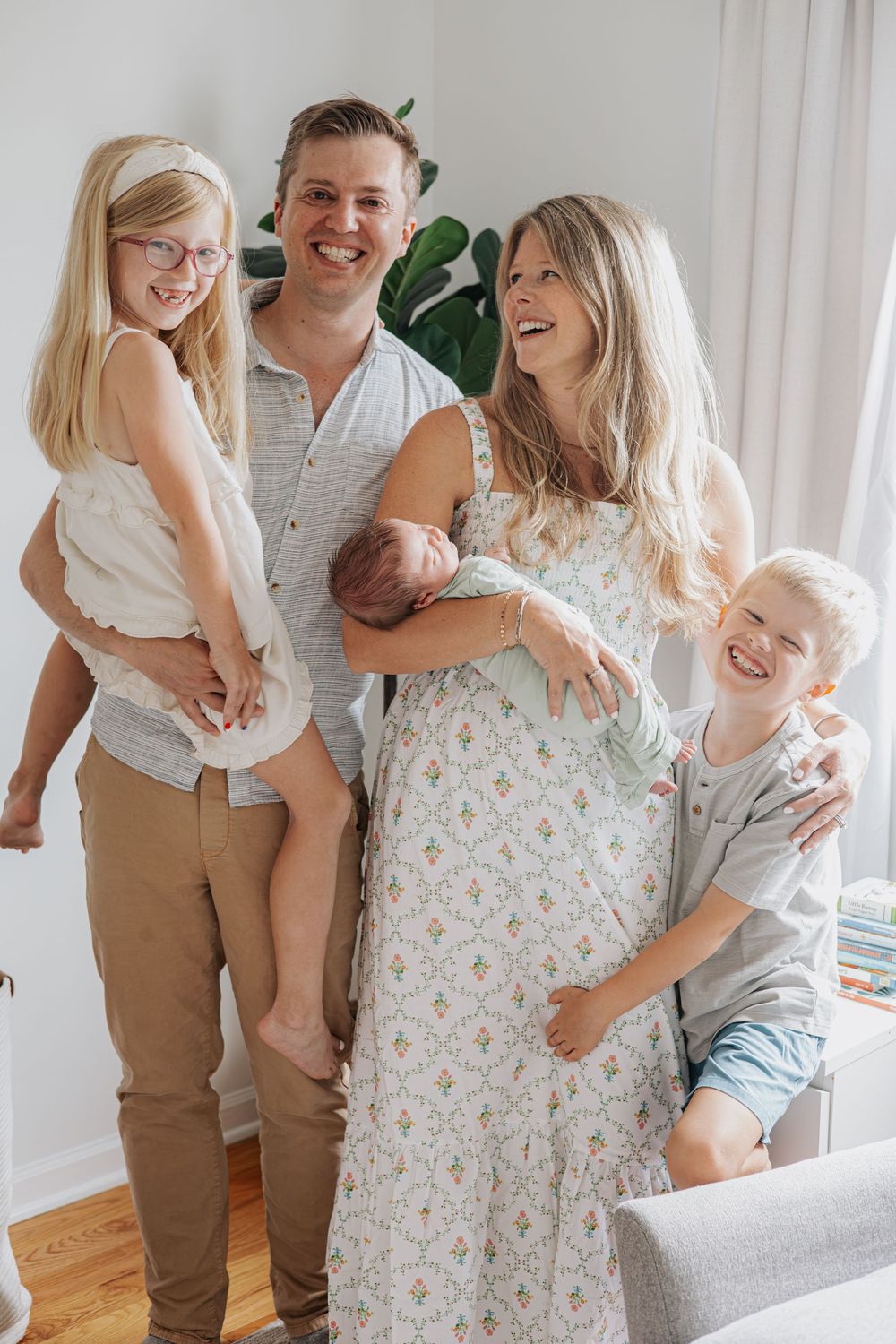 Happy family of five posing indoors, parents holding children including a newborn baby, smiling in a bright living room.