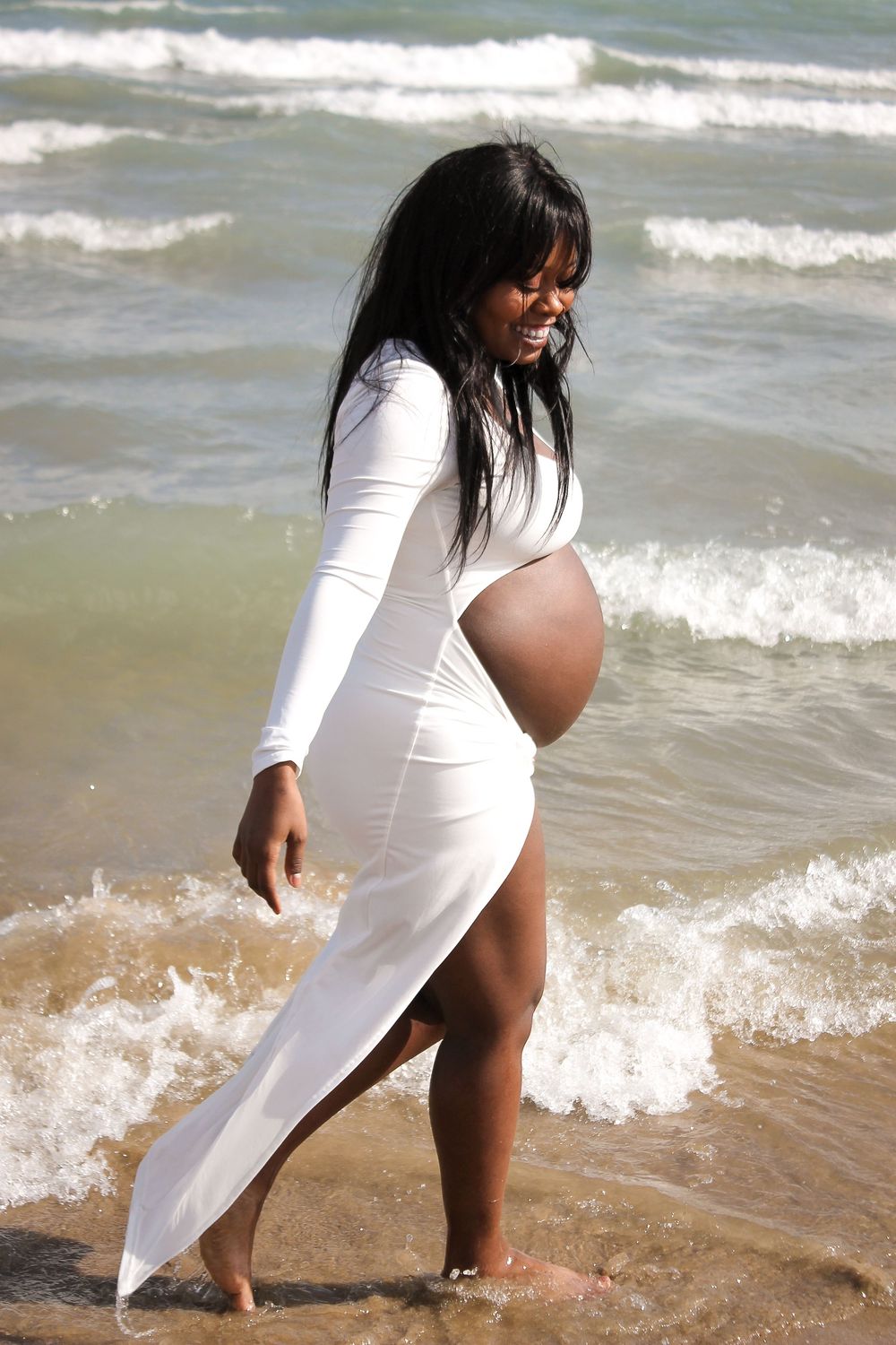 Person in white dress walking along beach shoreline during maternity photoshoot.