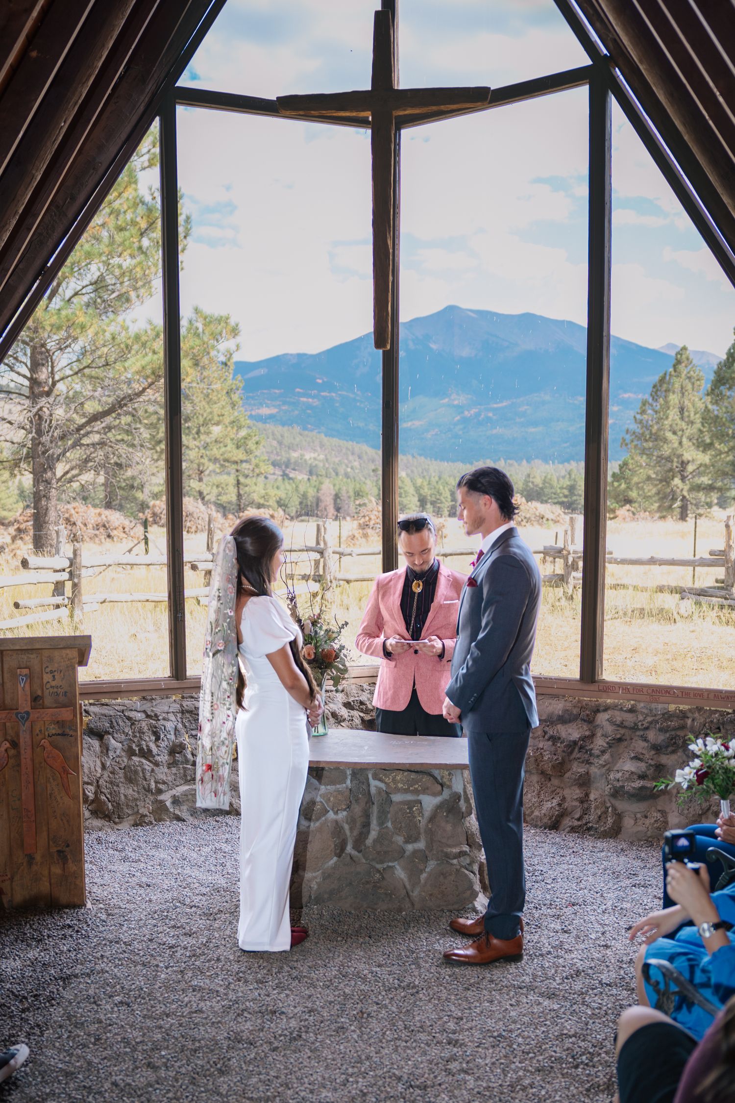 A wedding ceremony takes place inside a mountain chapel with scenic views of peaks through large windows.