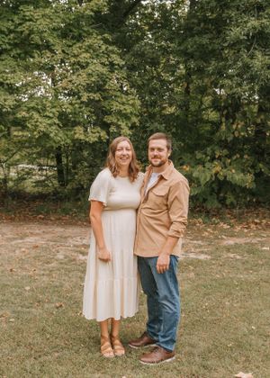 A couple stands together smiling in a park with fall foliage in the background.