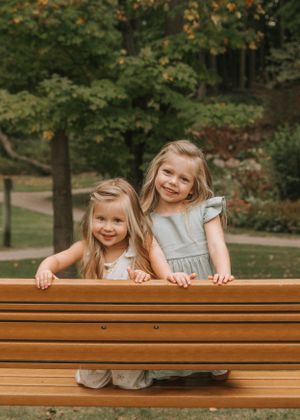 Two children peek over a wooden bench in a park with autumn trees in the background.