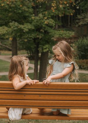 Two children play and interact while standing at a wooden bench in a park setting.