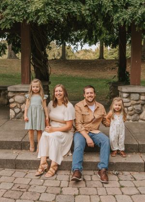 A family sits together on stone steps under a pergola in a park setting.