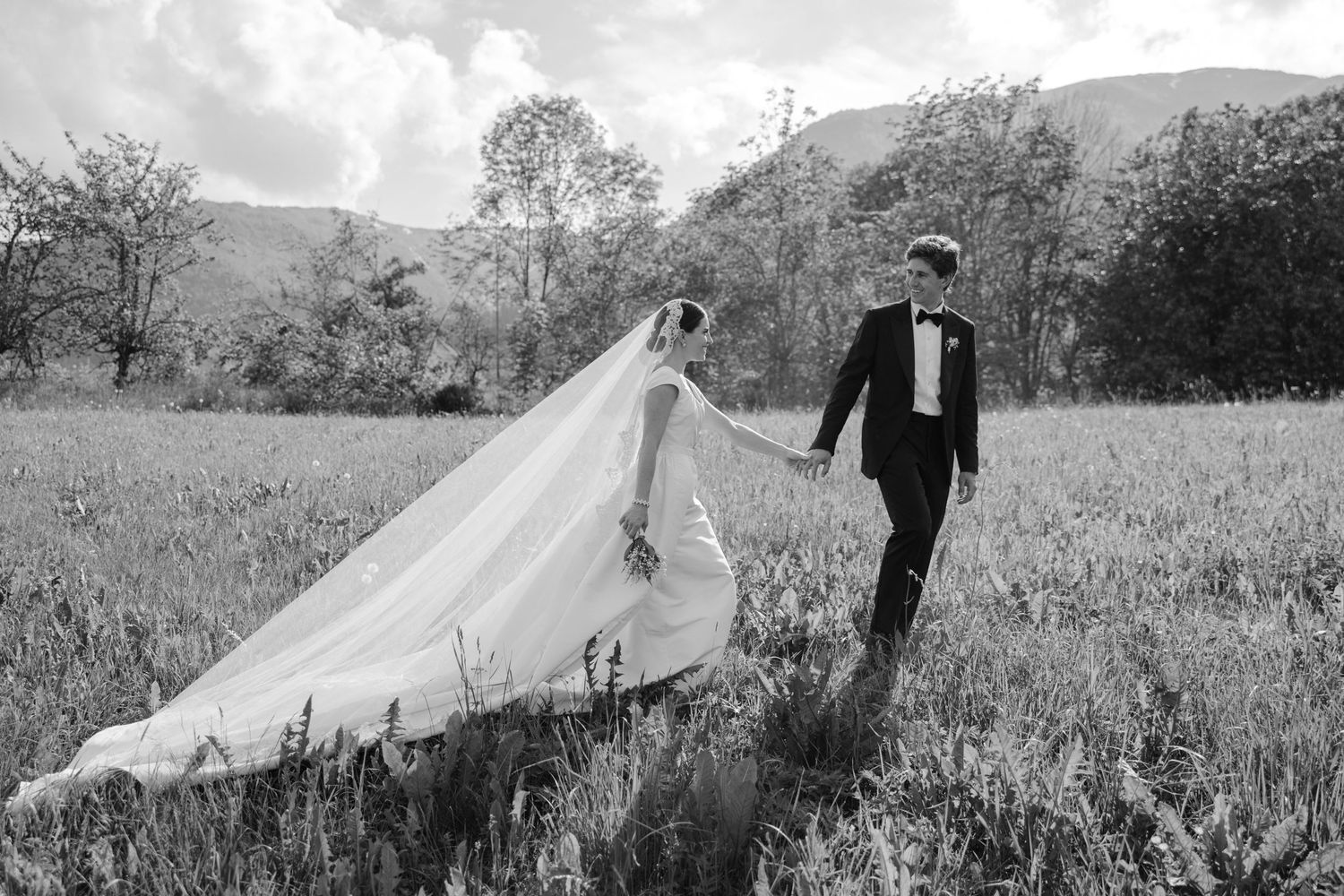 A romantic black and white photo of newlyweds walking through a meadow with flowing veil.
