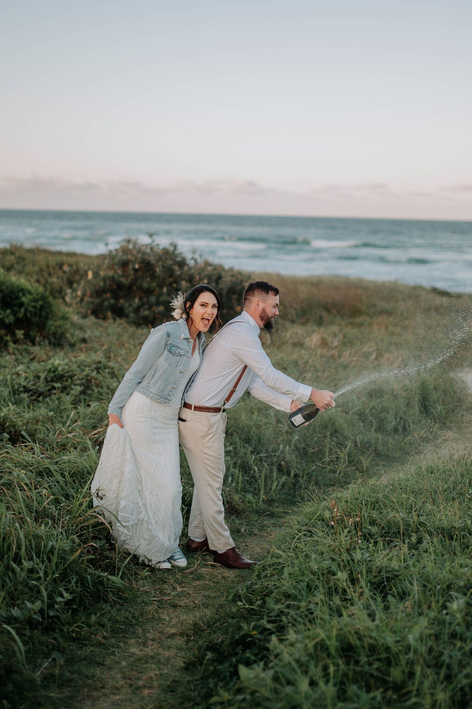 Playful wedding couple walking along coastal path in elegant attire.