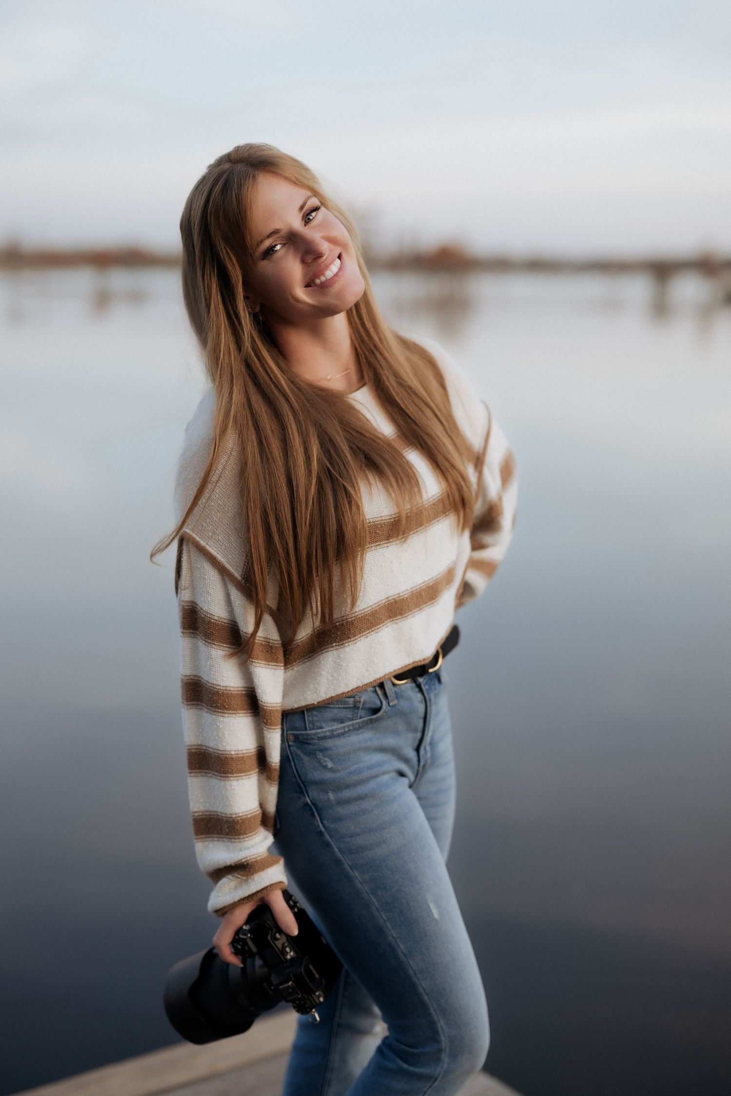 A person in a striped sweater and jeans poses playfully by a lake on an overcast day.