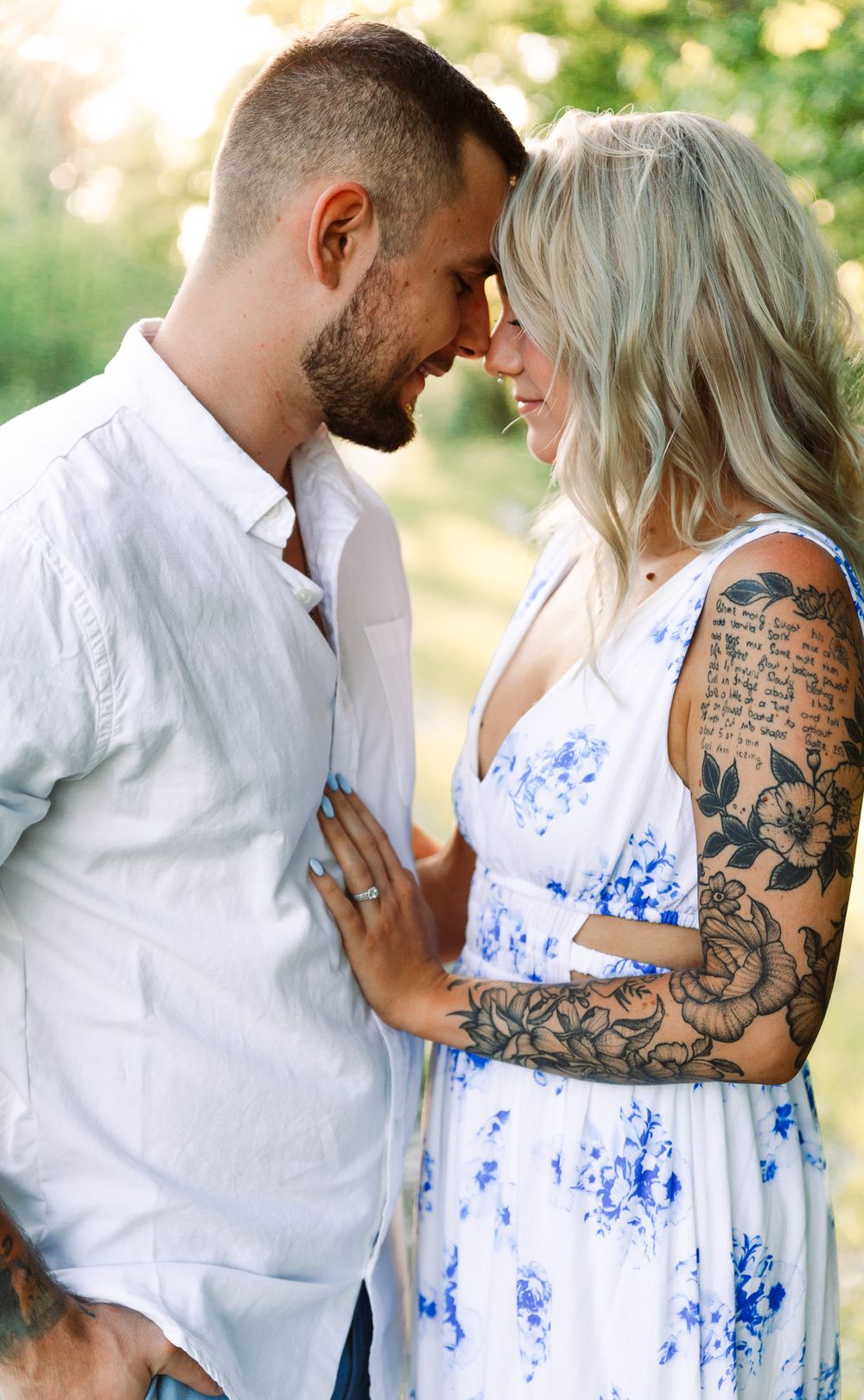 Romantic couple embraces outdoors, with one wearing a white shirt and tattoo sleeve and other in blue floral dress.