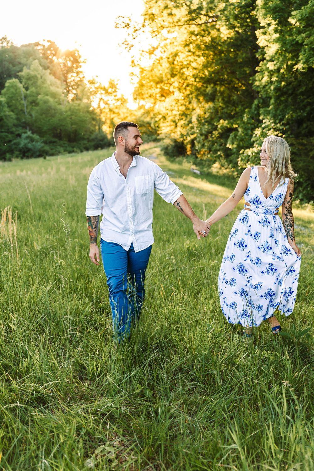 Couple walks hand in hand through golden-lit field with trees in background.