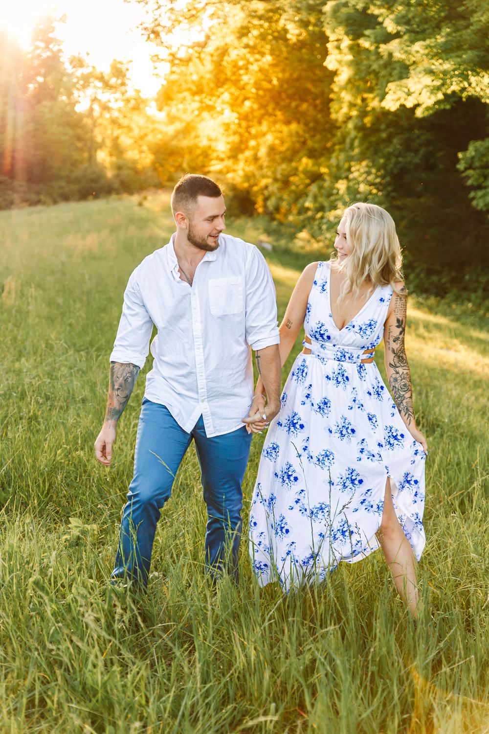 Couple strolls through sunlit meadow wearing white shirt and blue floral dress.