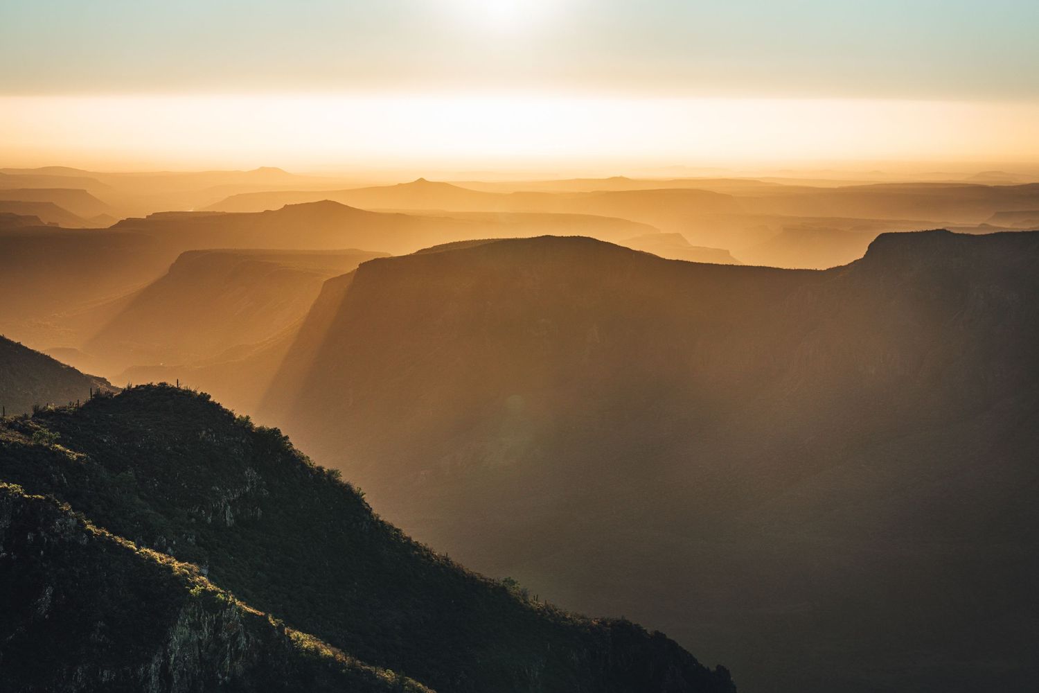 Scenic mountain landscape silhouettes at sunset with golden sunbeams streaming through layers of peaks in the distance.