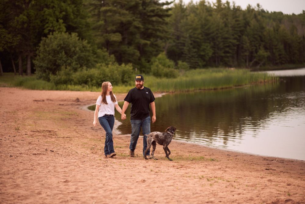 Couple walking hand in hand with a dog along a sandy lakeside beach surrounded by lush green trees.
