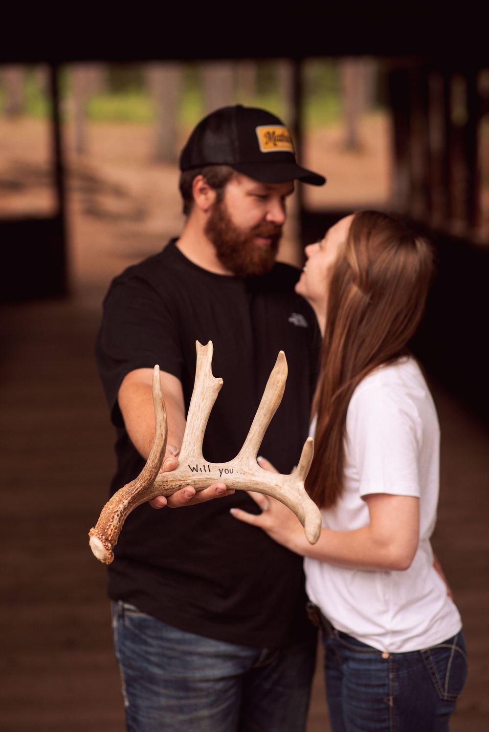 Couple holding deer antler with Will You written on it during outdoor proposal photo shoot.