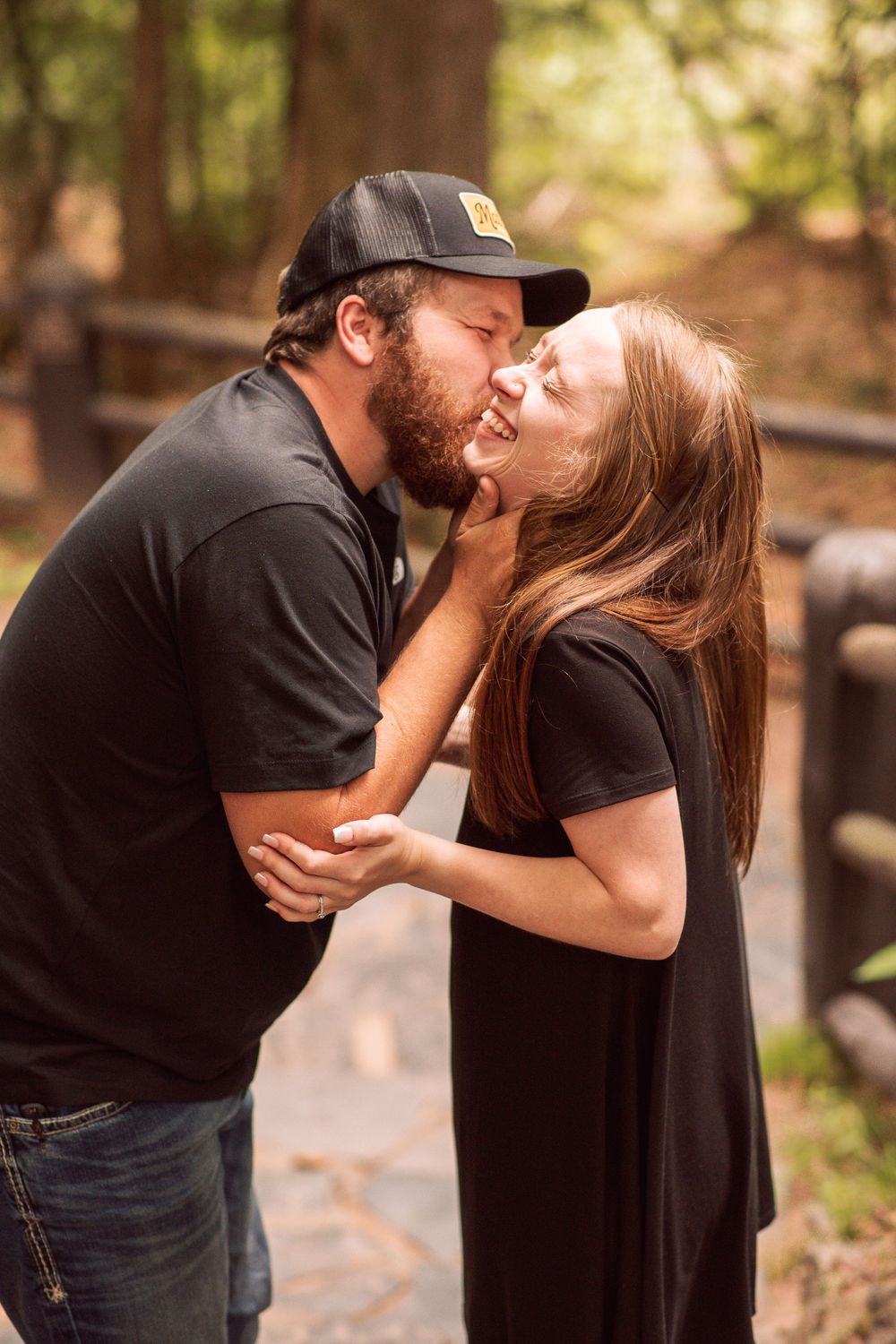 A bearded man in a black cap kisses a smiling woman in a black dress outdoors in a wooded setting.