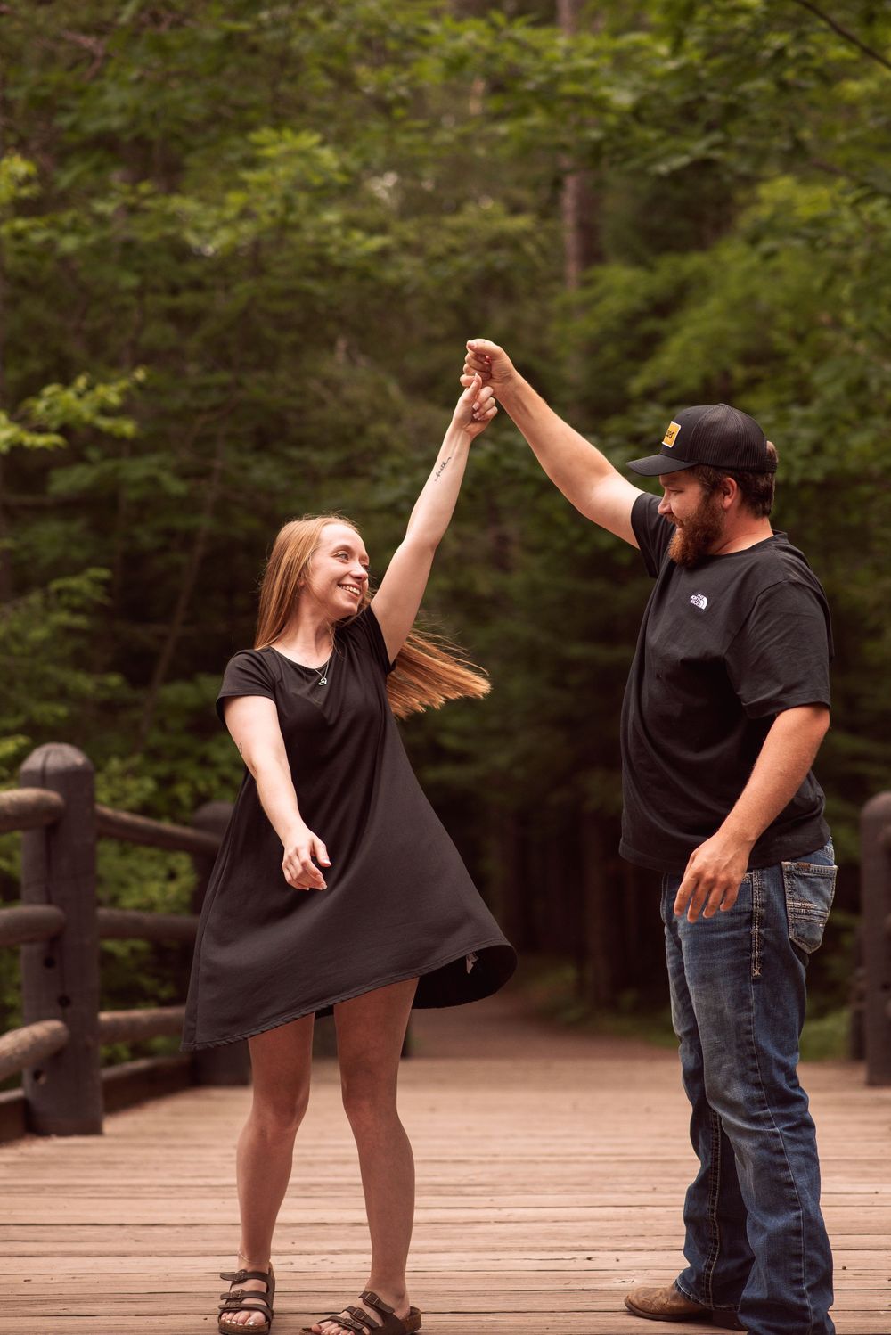 A couple dances joyfully on a wooden bridge surrounded by lush green forest, man spinning woman in black dress.