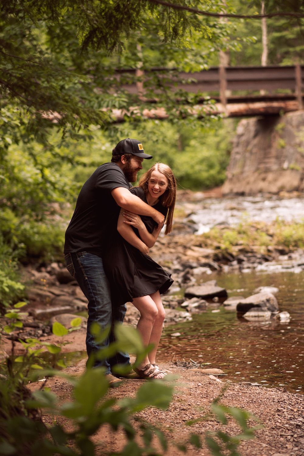 Couple laughing and embracing by a rocky creek in a lush green forest with a wooden bridge in the background.