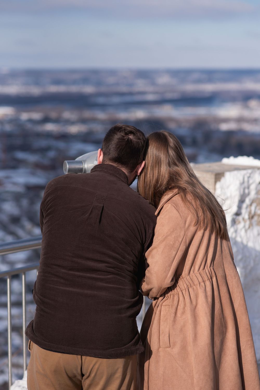 Couple sharing a coin-operated telescope at a snowy scenic overlook on a sunny winter day.