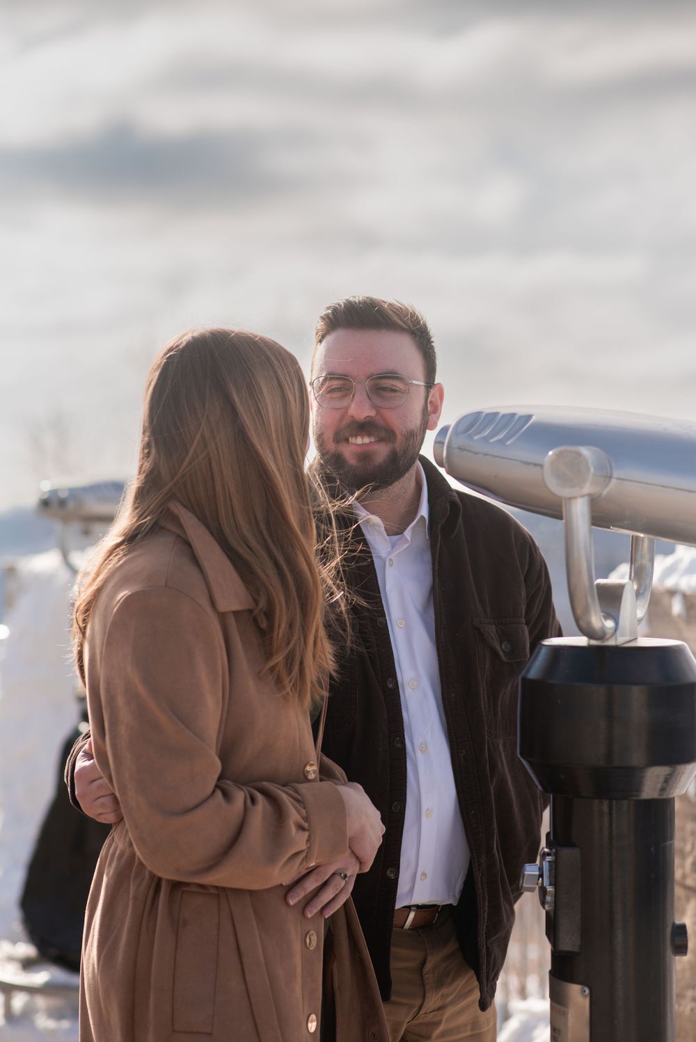 Couple smiling at each other outdoors in winter, man with glasses and beard, woman in brown coat.