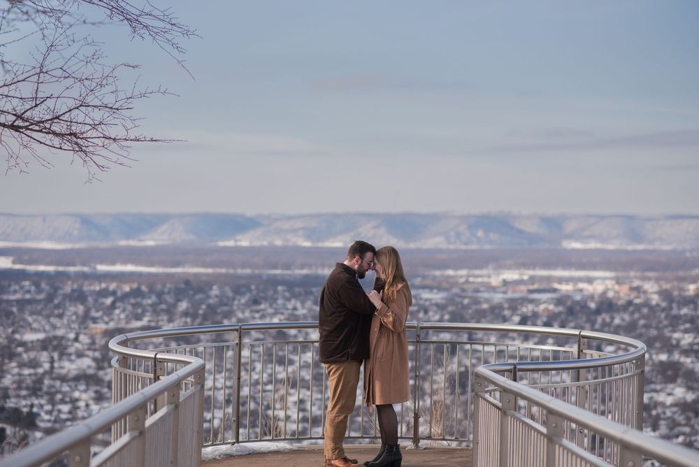 Couple embracing on a scenic overlook with snowy cityscape and mountain views on a clear winter day.